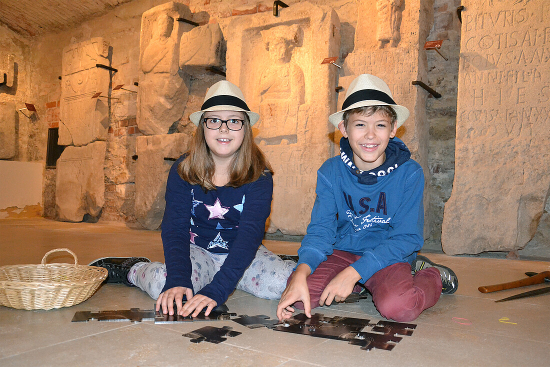 Ein Mädchen und ein Junge sitzen vor einer archäologischen Sammlung