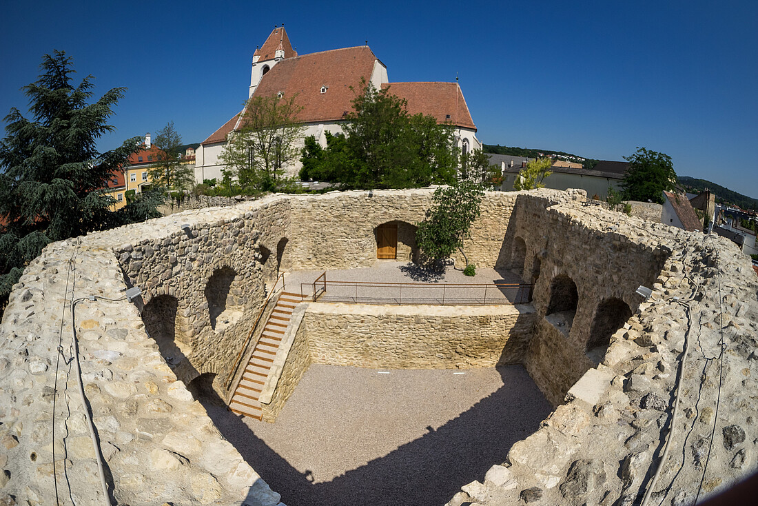 Runder, offener alter Pulverturm aus groben Steinen errichtet. Im Hintergrund eine Kirche.